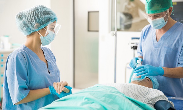 Two healthcare professionals wearing masks, gloves and scrubs care for a patient in a hospital bed.